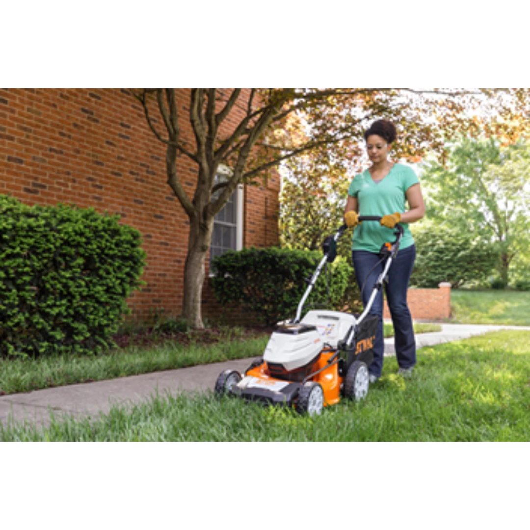 Woman using orange and white self-propelled battery lawn mower on grass near brick building in outdoor power equipment setting