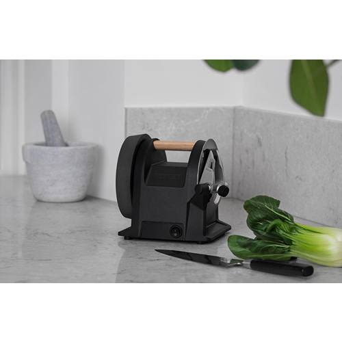 Compact carbon black kitchen knife sharpener with wooden handle on marble countertop next to cutting knife and greens