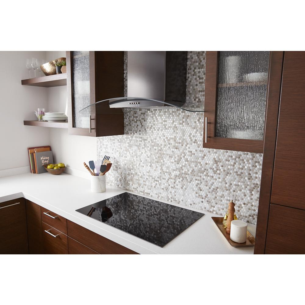 Kitchen countertop with black electric cooktop and wall-mounted concave glass range hood in modern wood cabinetry setting