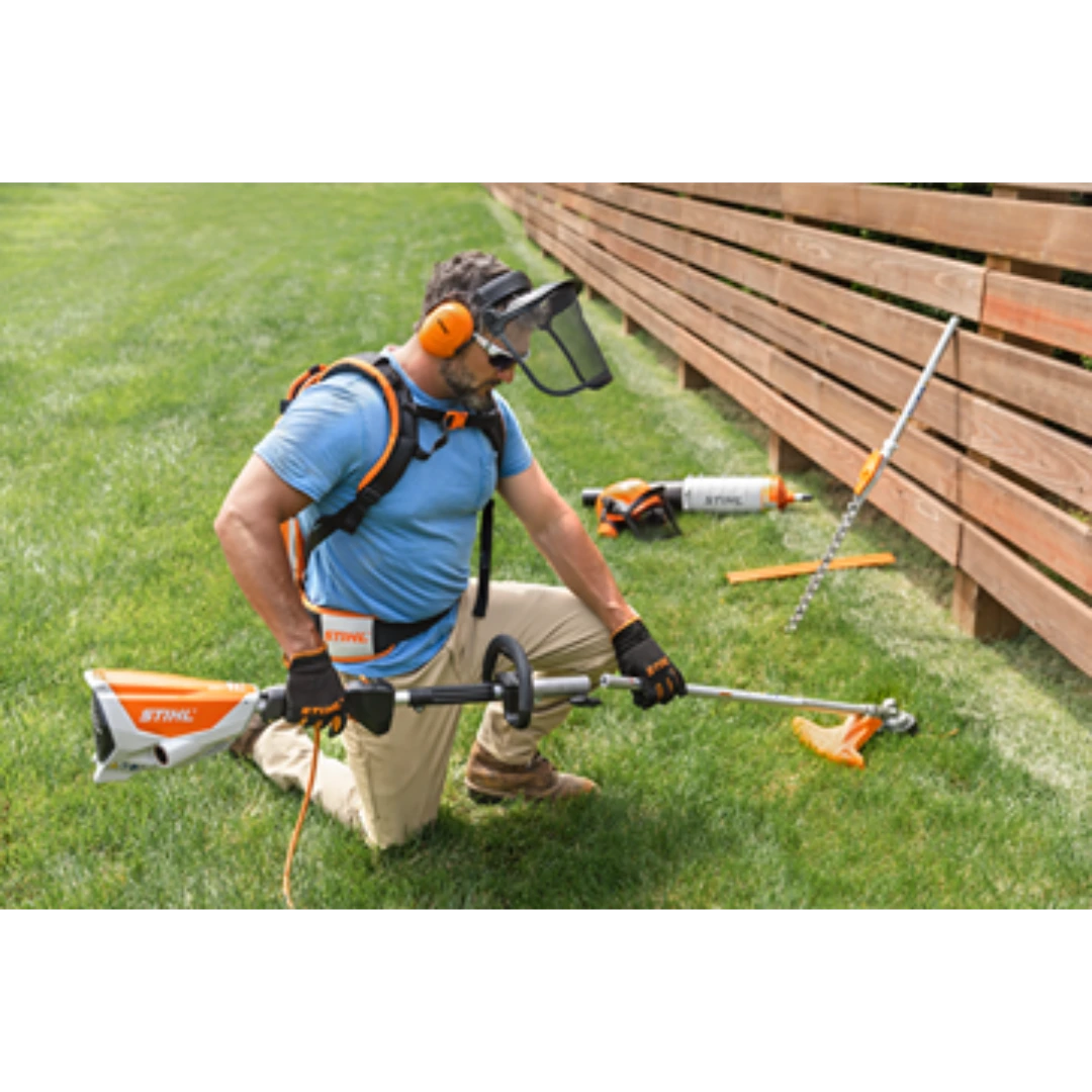 Man wearing protective gear using a battery-powered trimmer near wooden fence in outdoor power equipment setting