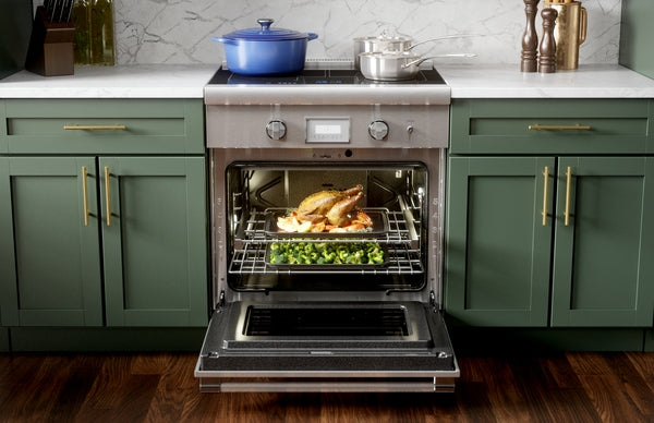Open stainless steel induction professional range with roasted chicken and vegetables inside, surrounded by green cabinetry and countertop
