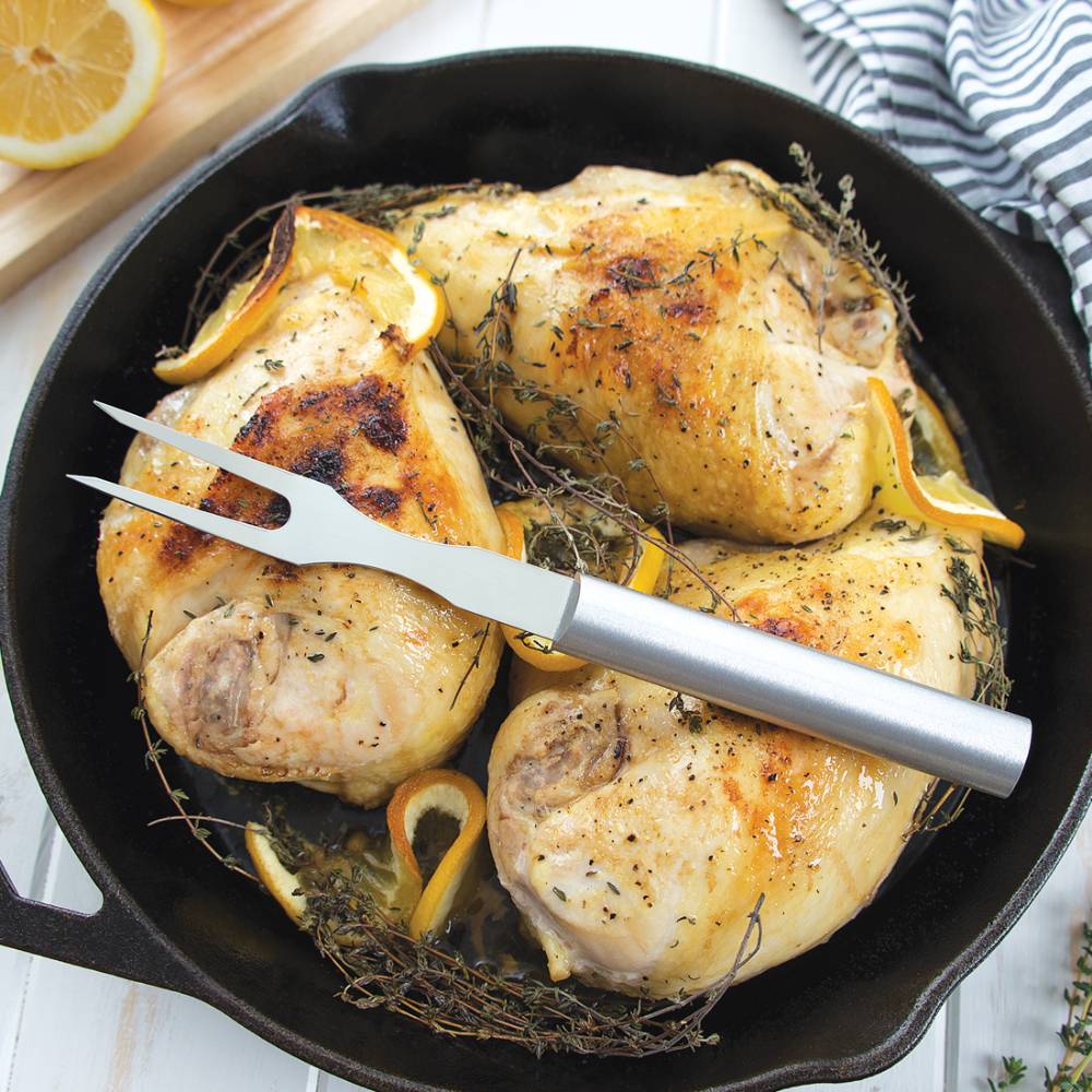 Two-pronged carving fork with slim handle resting on roasted chicken with lemon and herbs in cast iron skillet in kitchenware collections