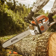 Person wearing gloves and plaid shirt operating an orange and white chainsaw cutting through a thick tree trunk outdoors in a wooded area