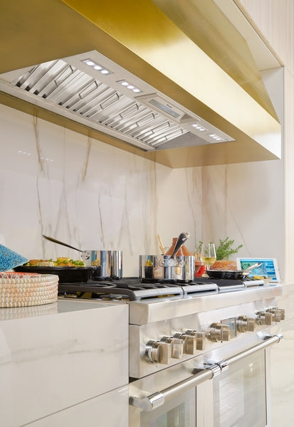 Stainless steel custom insert range hood above a modern stove in a kitchen with marble backsplash and gold trim