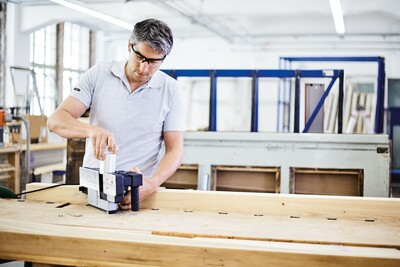 Man using a Festool electric planer on a large wooden board in a well-lit workshop for home improvement tasks