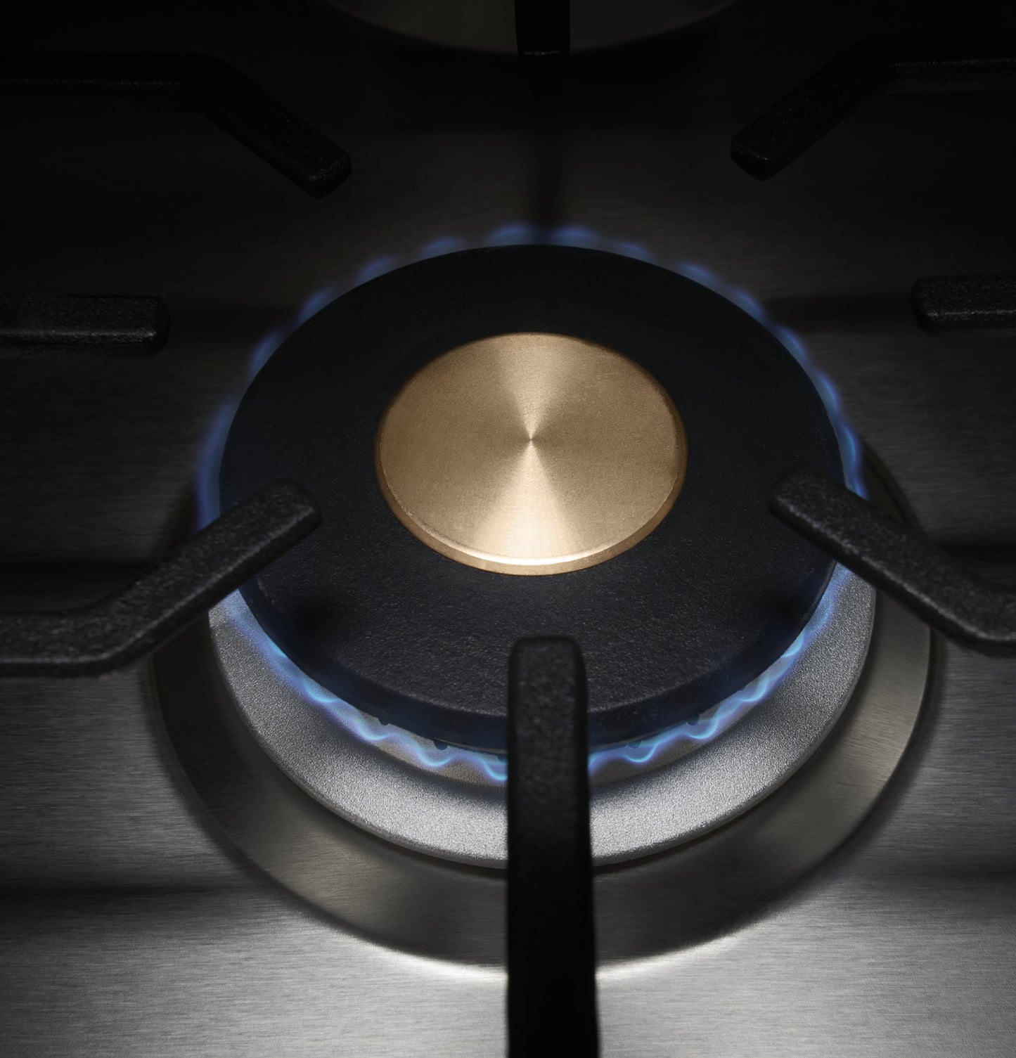 Close-up view of brass burner with blue flame and cast iron grate on stainless steel cooktop for kitchenware and appliances.