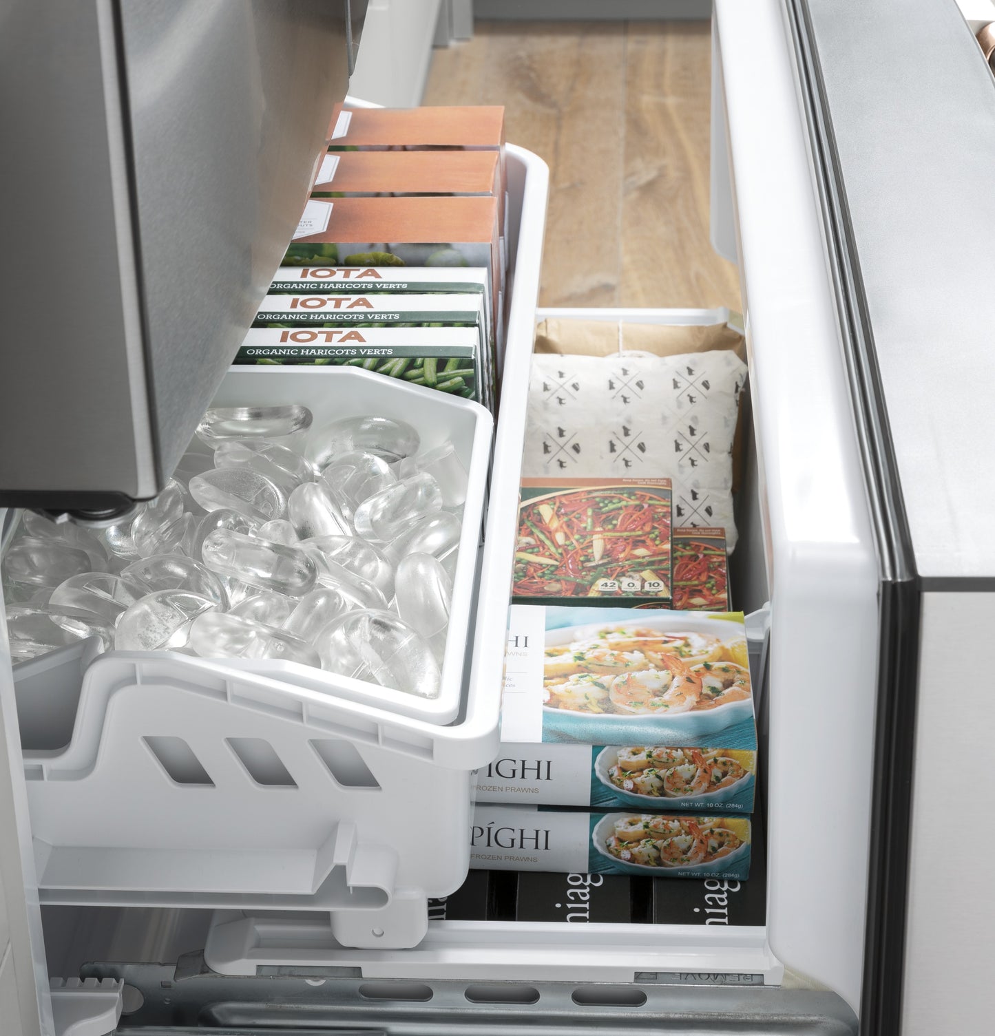 Open freezer drawer showing clear ice bin, frozen meals, and packaged food inside a stainless steel refrigerator