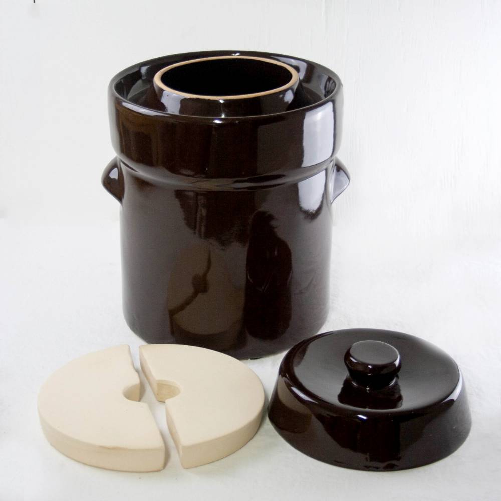 Glossy dark brown ceramic fermenting crock with separate lid and two white airlock weights on white background