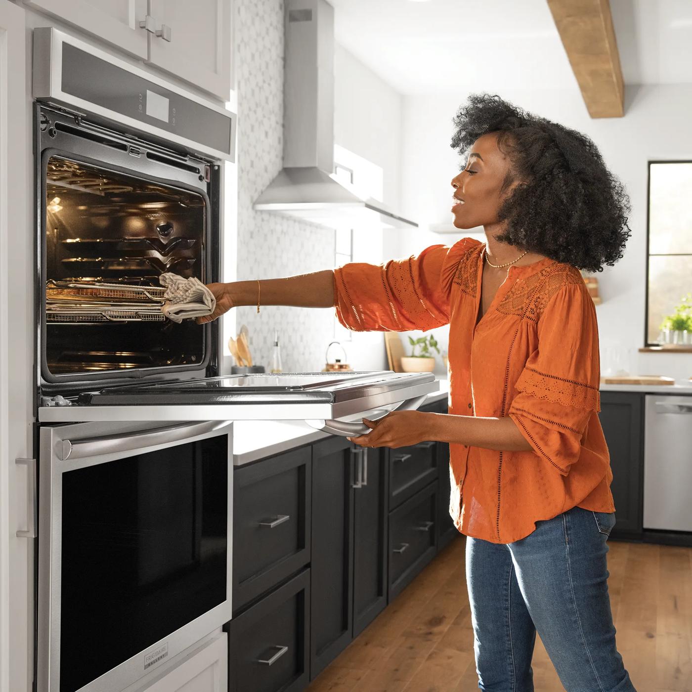 Woman wearing orange shirt placing pan into double wall oven in modern kitchen with black and white cabinetry