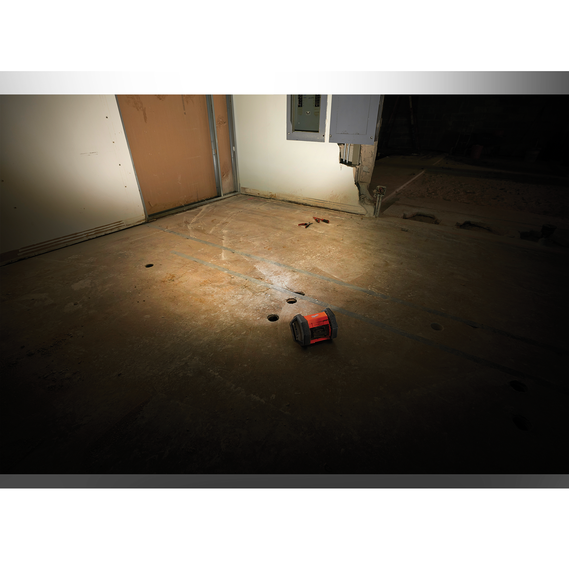 Compact Milwaukee red and black work light illuminating concrete floor near wall panels in utility space Tools collection
