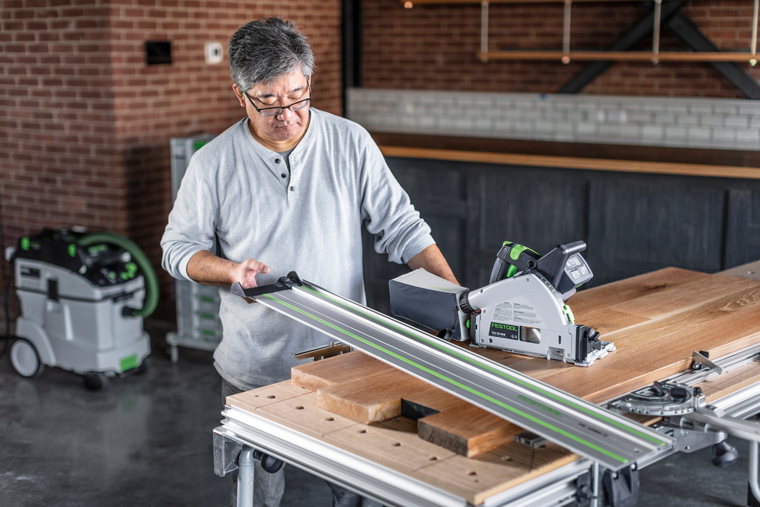 Man using cordless Festool track saw with guide rail to cut wood on workbench in workshop for home improvement task