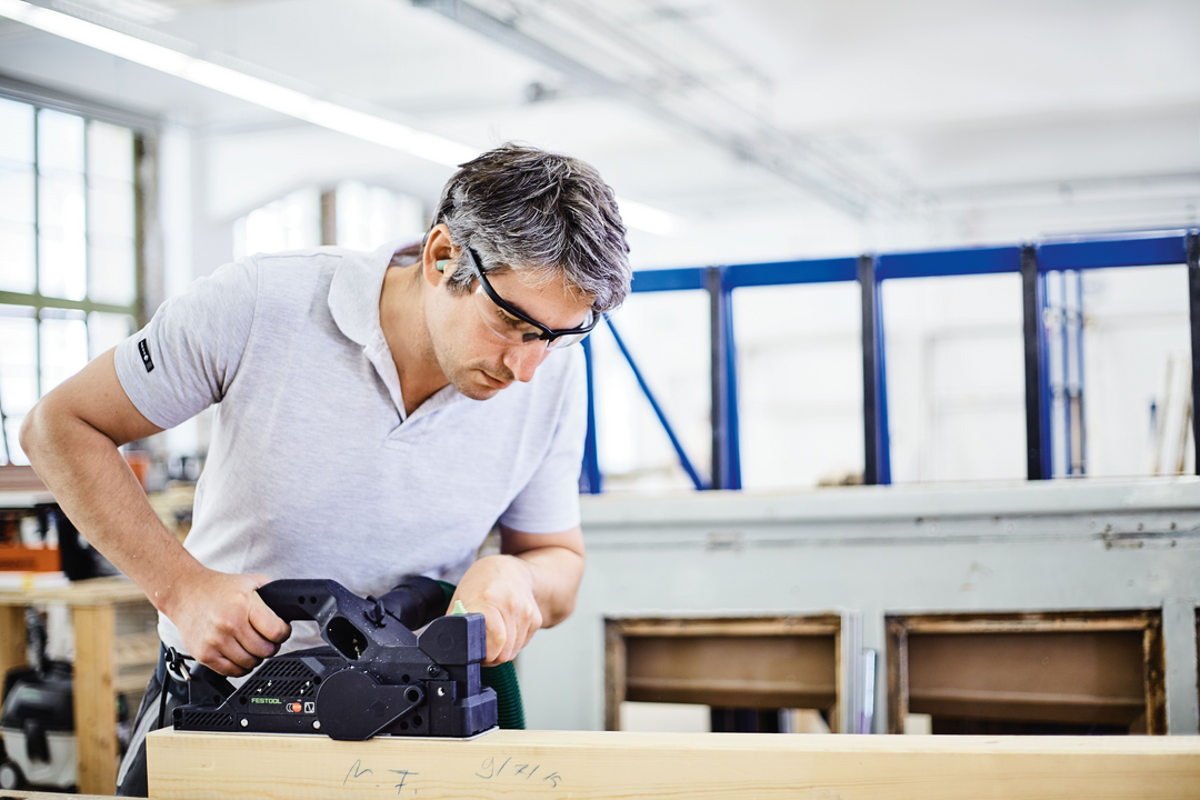 Man wearing safety glasses using an electric planer on wood in a bright workshop for home improvement tasks