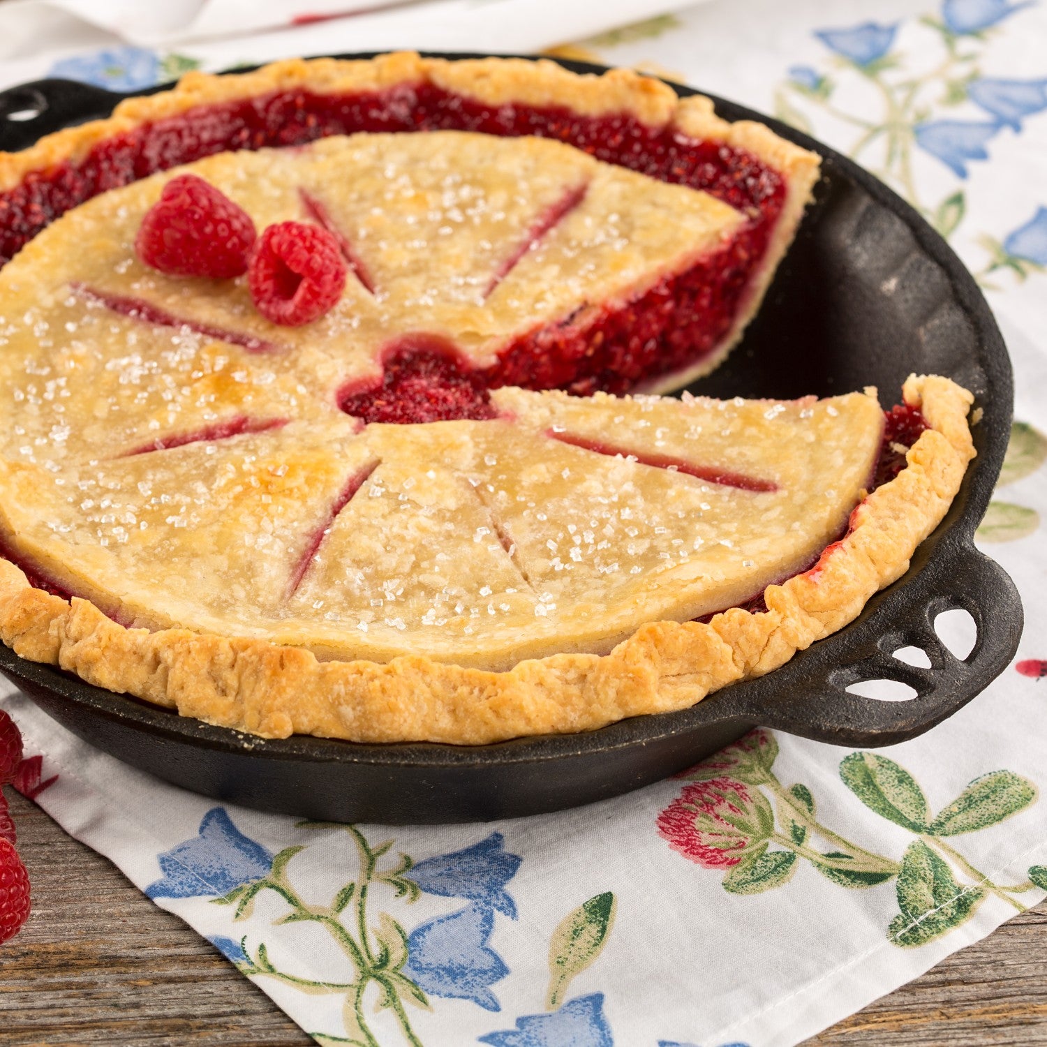 Fresh raspberry pie with golden crust in cast iron pan with crimped edges on floral cloth in kitchenware collection