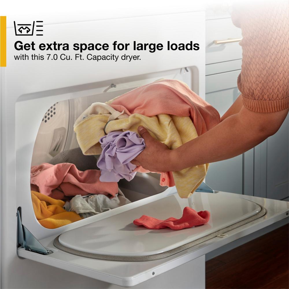 Person placing colorful clothes into open white top load gas dryer in a home laundry area with gray cabinetry and copper handle