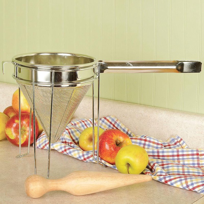 Stainless steel food mill with fine mesh and handle on kitchen counter with apples and wooden tamper