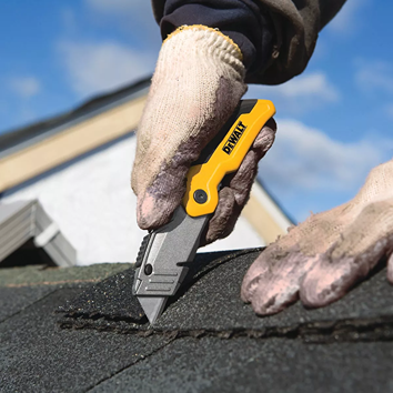 Hand in glove using a compact yellow and gray retractable utility knife cutting roofing shingles in home improvement tools collection