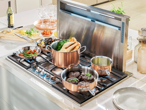 Stainless steel 36-inch gas cooktop with copper cookware cooking vegetables, pasta, and beef patties in modern kitchen setting
