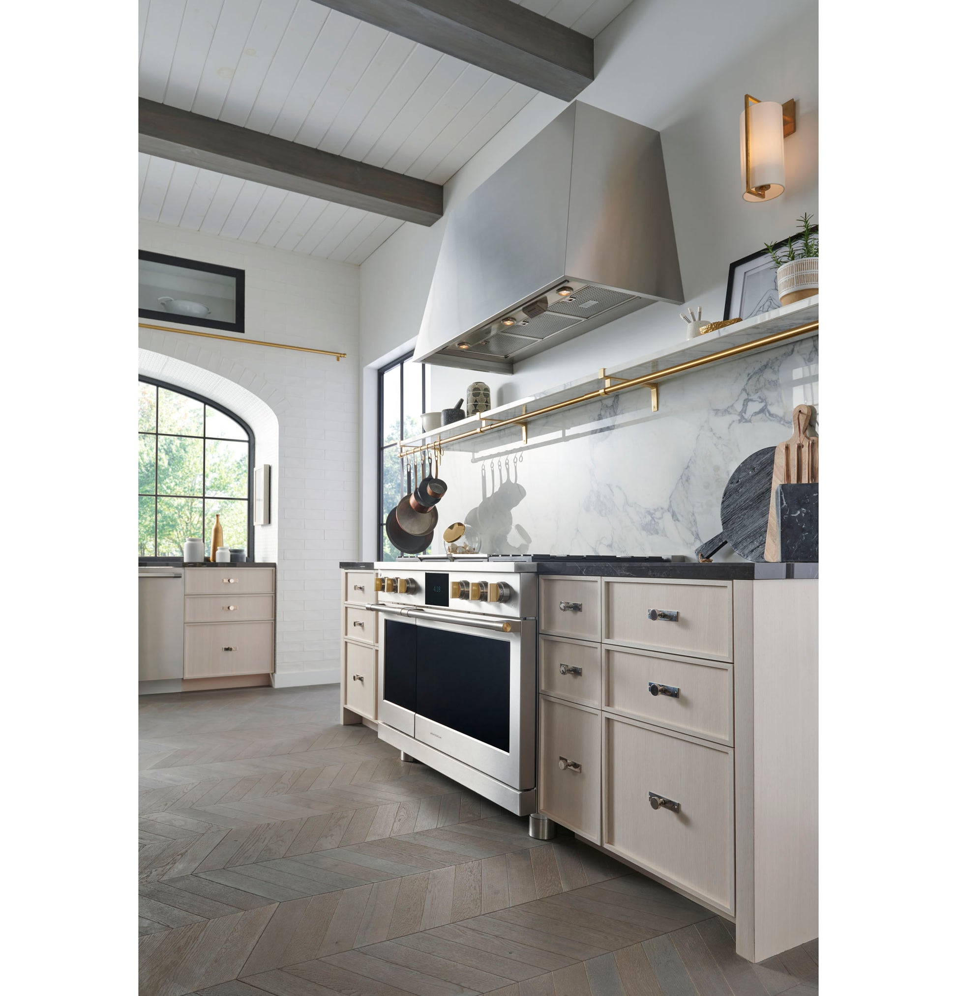 Modern kitchen with stainless steel range hood, light cabinetry, marble backsplash, and brass rail for cookware hanging