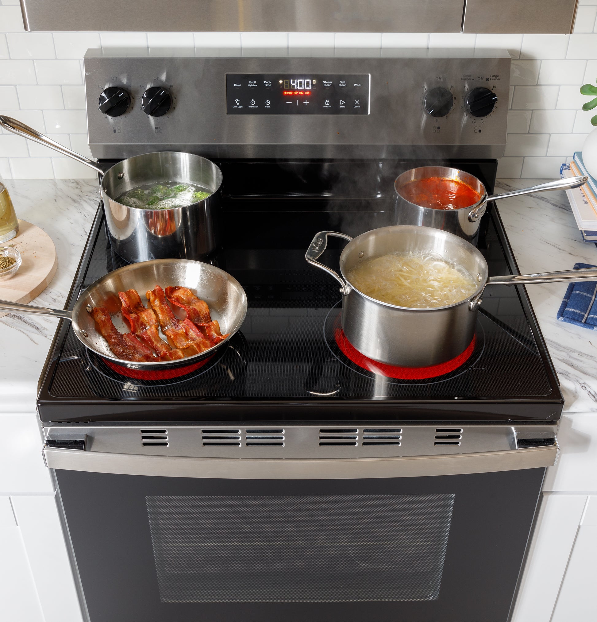 Free-standing electric range with four pots and pans cooking bacon, pasta, sauce, and boiling vegetables