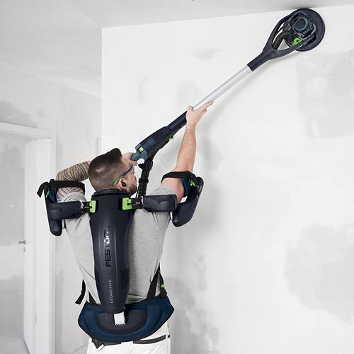 Man wearing blue and black ergonomic exoskeleton using a long-handled power sander on an indoor wall surface