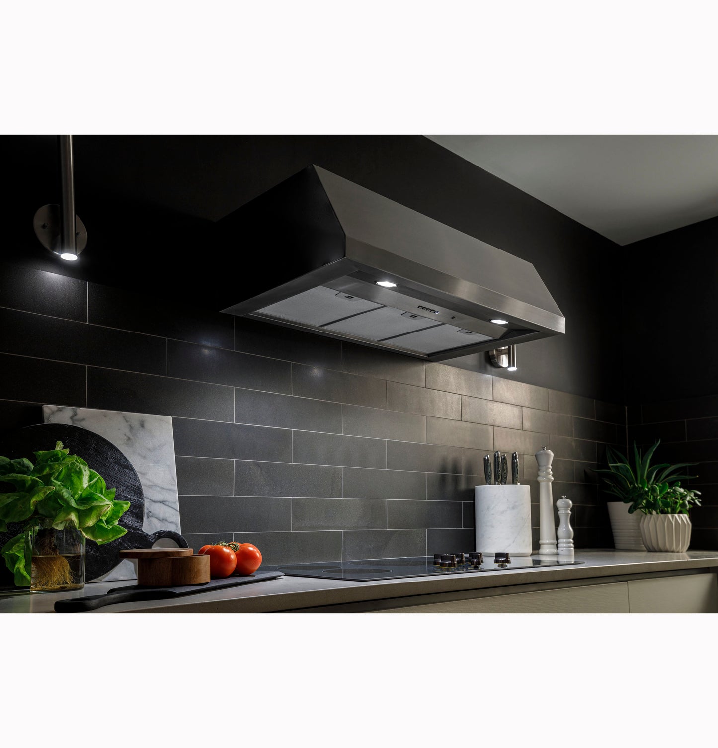 Stainless steel kitchen hood with built-in lights over modern stovetop in a sleek, dark tile backsplash setting.
