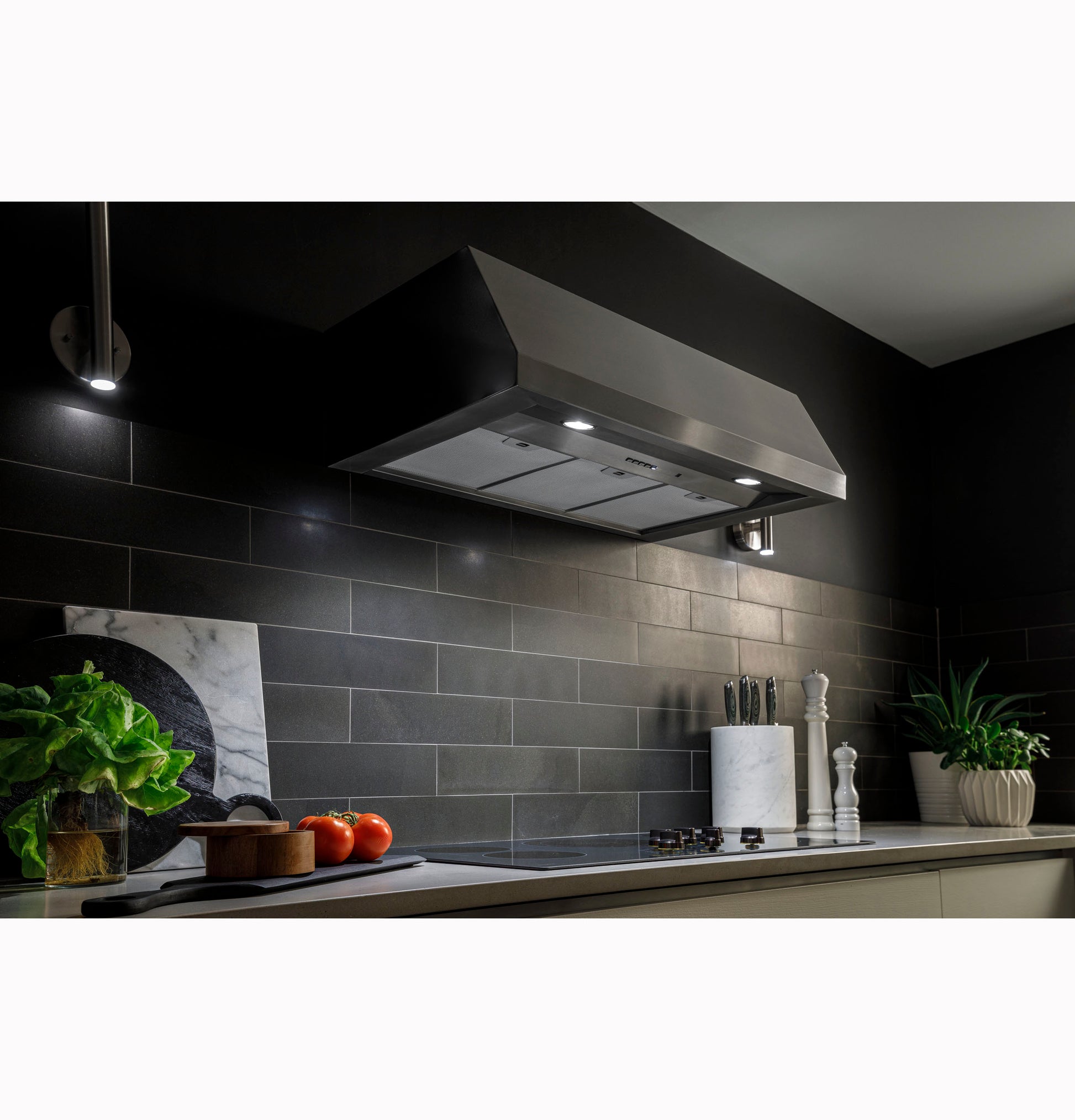 Stainless steel kitchen hood with built-in lights over modern stovetop in a sleek, dark tile backsplash setting.