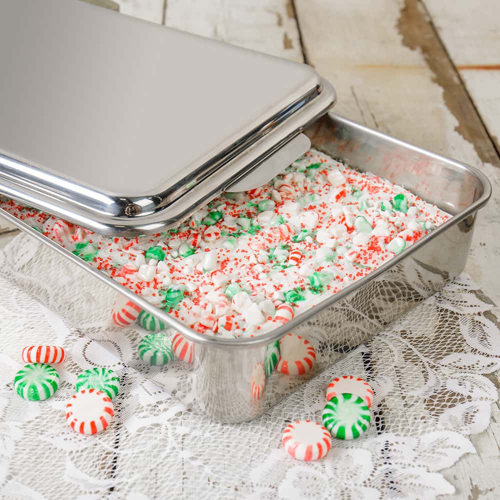 Rectangular stainless steel cake pan with partially removed lid, filled with festive dessert and scattered peppermint candies