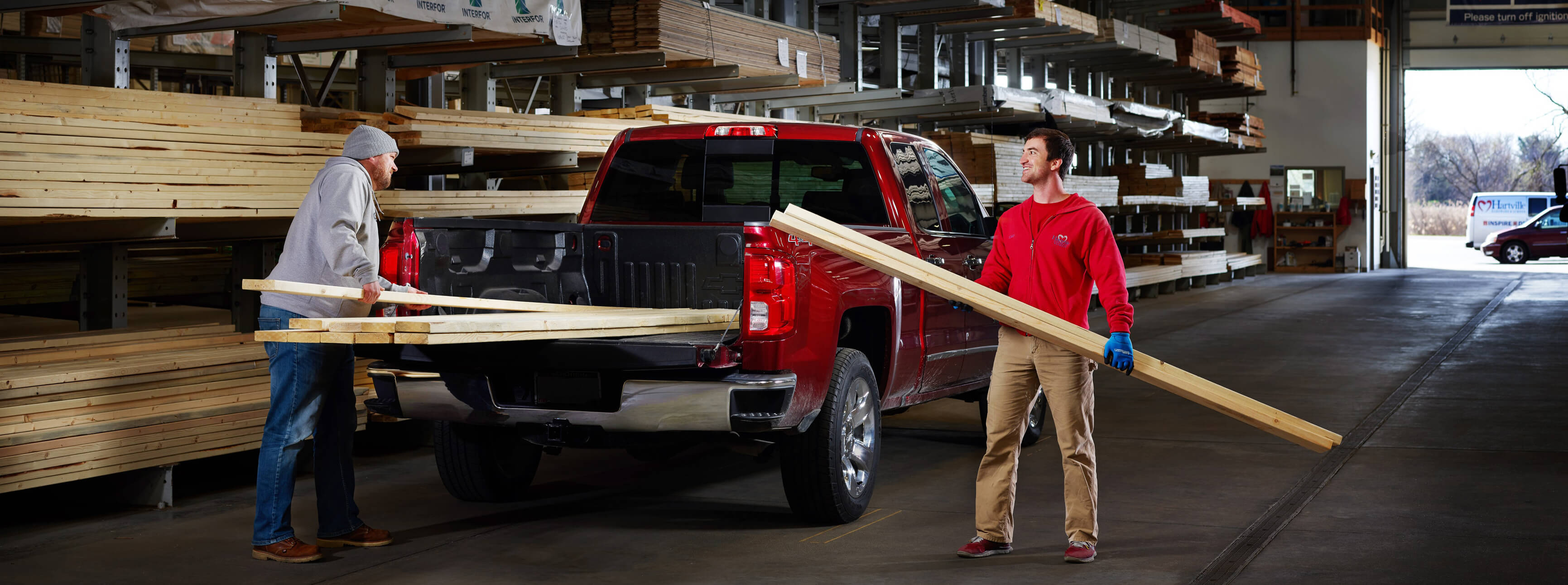 Two men loading long wooden planks into the bed of a red pickup truck inside a large lumber warehouse with shelves stocked with various wood materials.