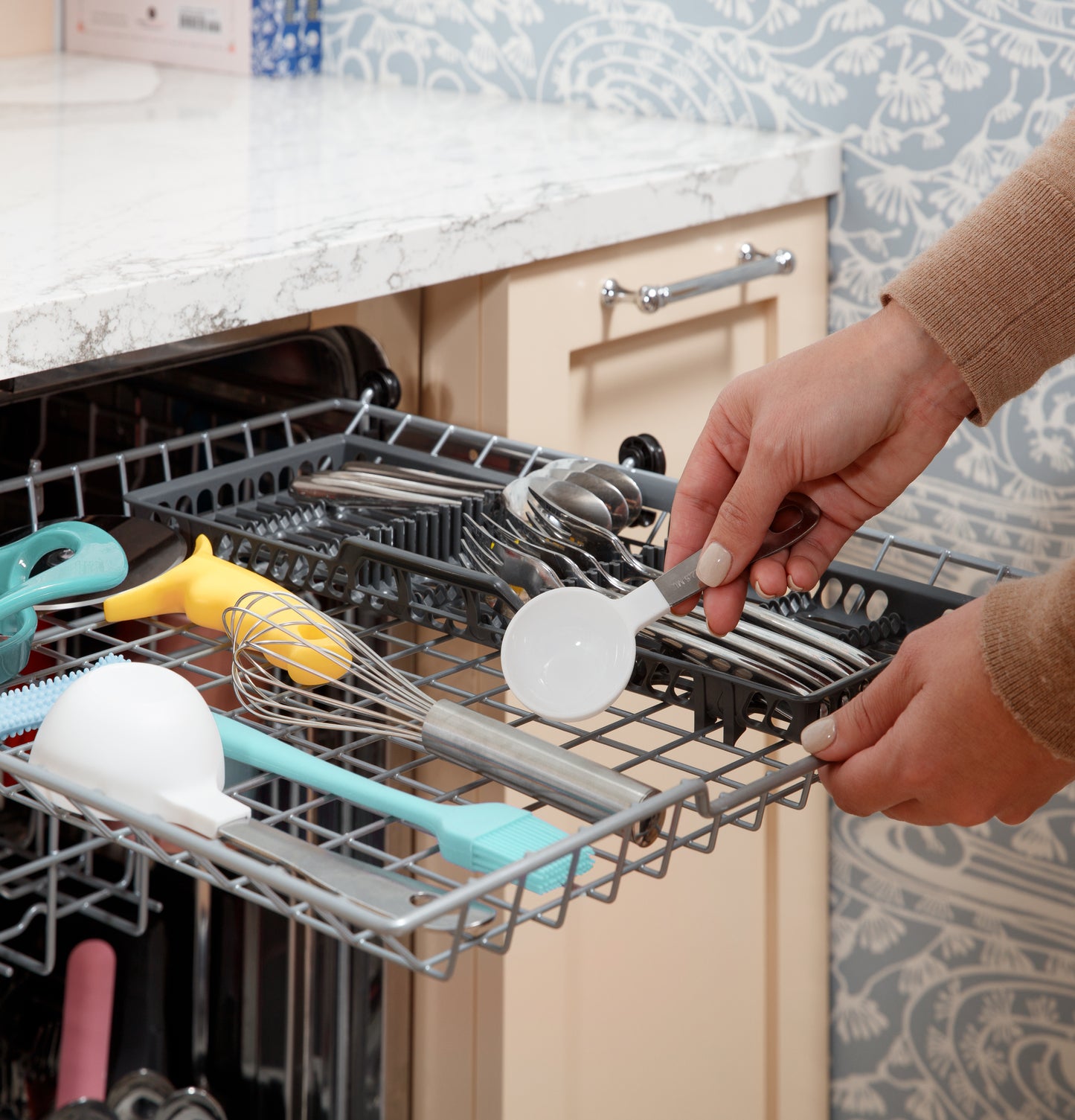Person placing a measuring spoon into the top rack of a stainless steel interior dishwasher in a kitchen setting