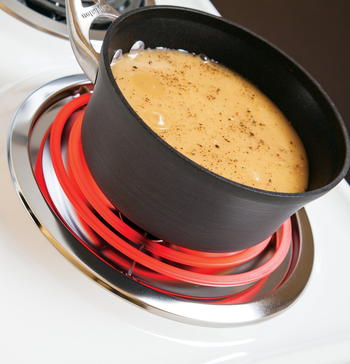 Black saucepan heating creamy sauce on glowing red coil burner of white electric range cooktop in kitchen appliance setting