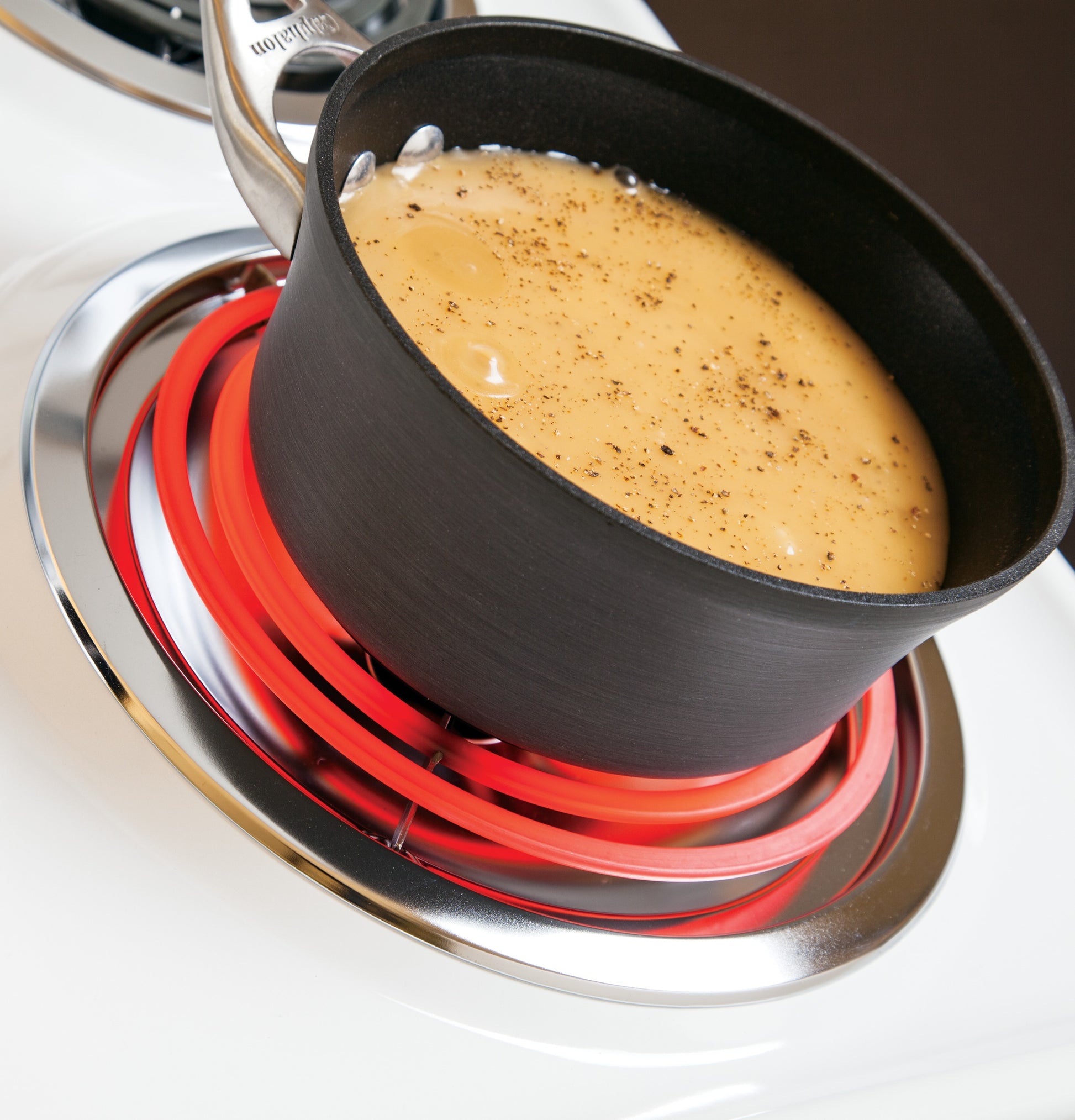 Black saucepan heating creamy sauce on glowing red coil burner of white electric range cooktop in kitchen appliance setting