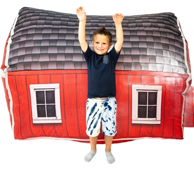 Child standing with arms raised in front of a red inflatable barn playhouse with printed windows and gray roof