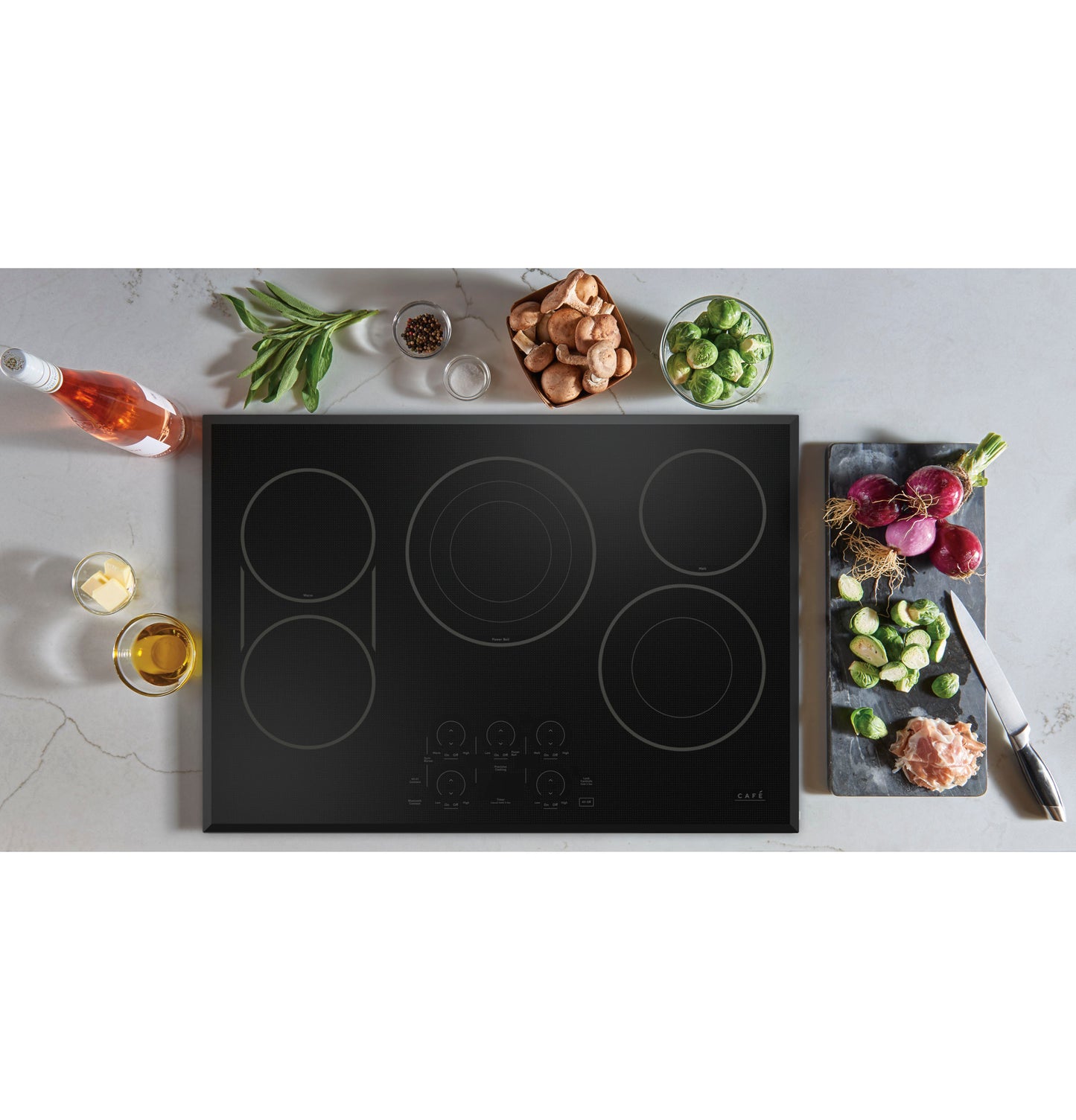 Black glass electric cooktop with touch controls surrounded by fresh vegetables and cooking ingredients on countertop