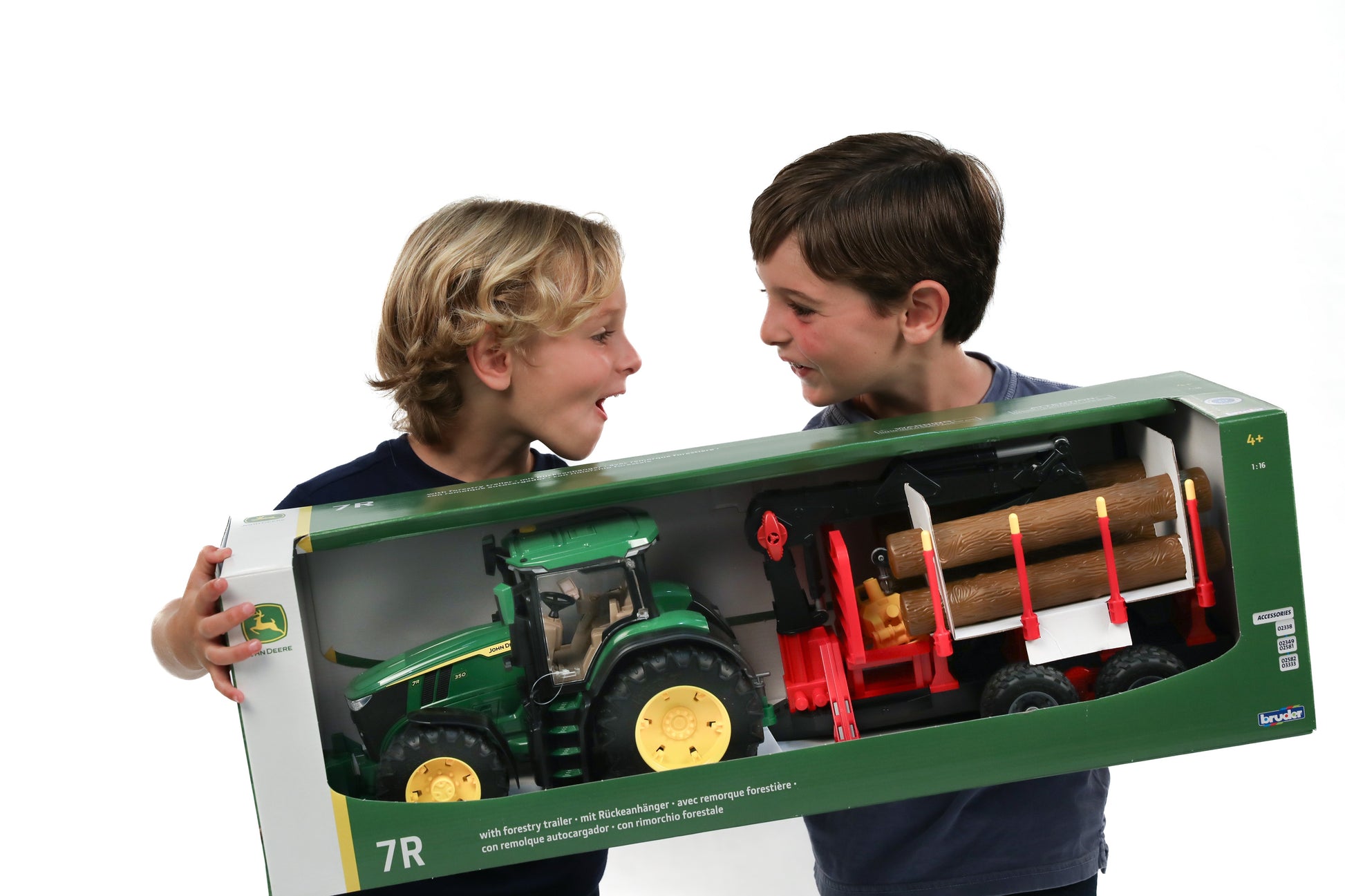 Two children holding and smiling at a green John Deere toy tractor with forestry trailer carrying wooden logs