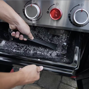 Person scraping ashes from a stainless steel gas grill’s firebox with control knobs and tool for cleaning