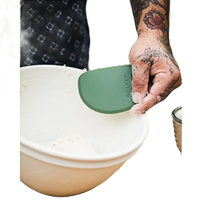 Green dough scraper with ergonomic design held over white mixing bowl for baking and food preparation tasks