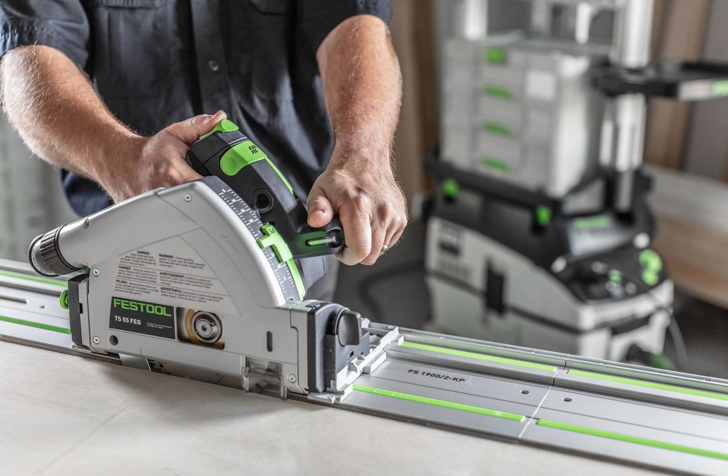 Person operating Festool plunge-cut track saw on guide rail on workbench in workshop with tools nearby