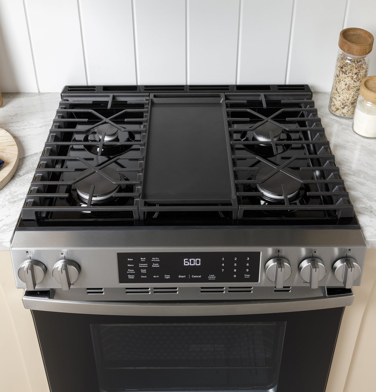 Stainless steel gas range with four burners and central griddle, digital display, and front control knobs in kitchen setting