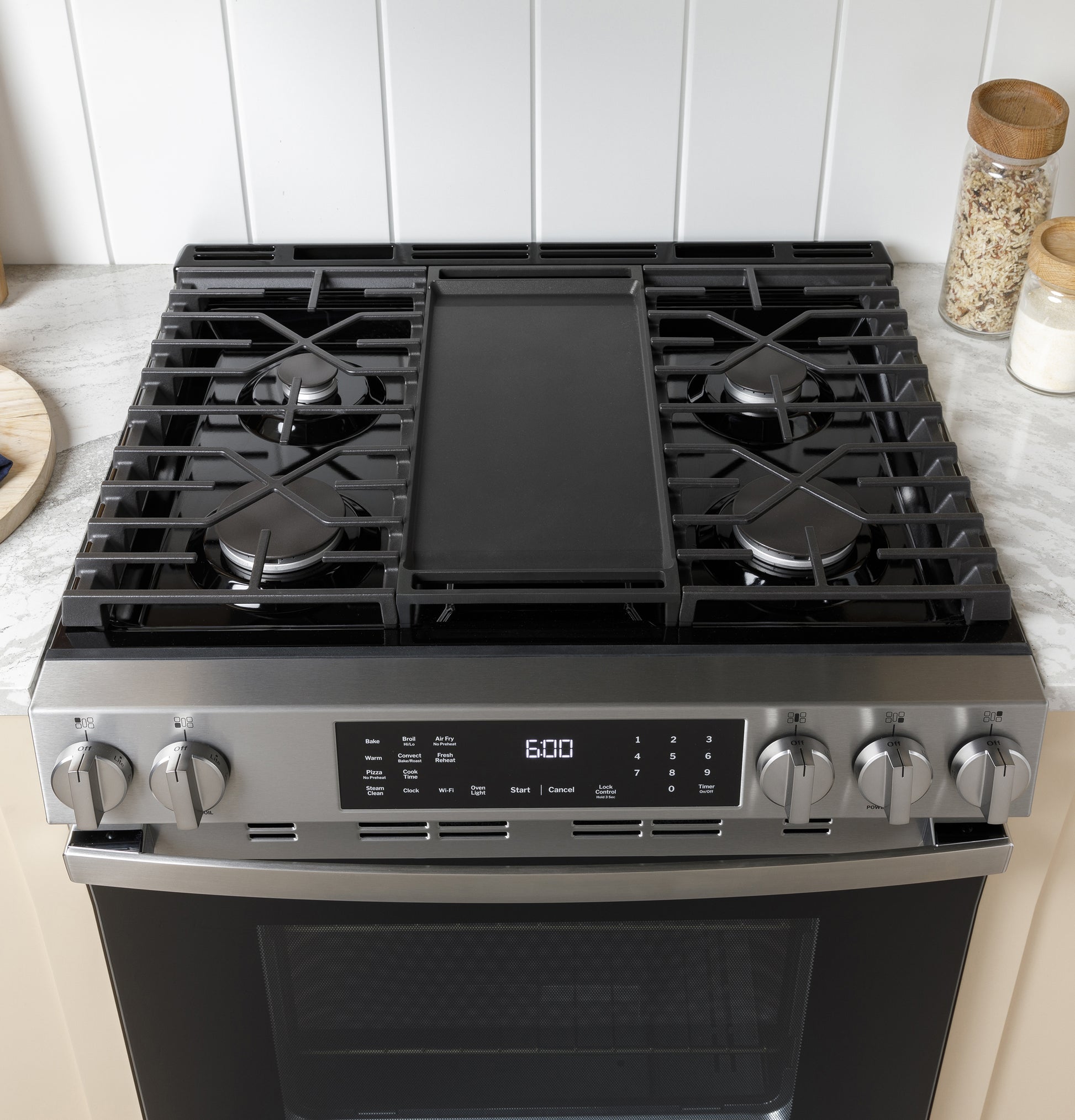 Stainless steel gas range with four burners and central griddle, digital display, and front control knobs in kitchen setting