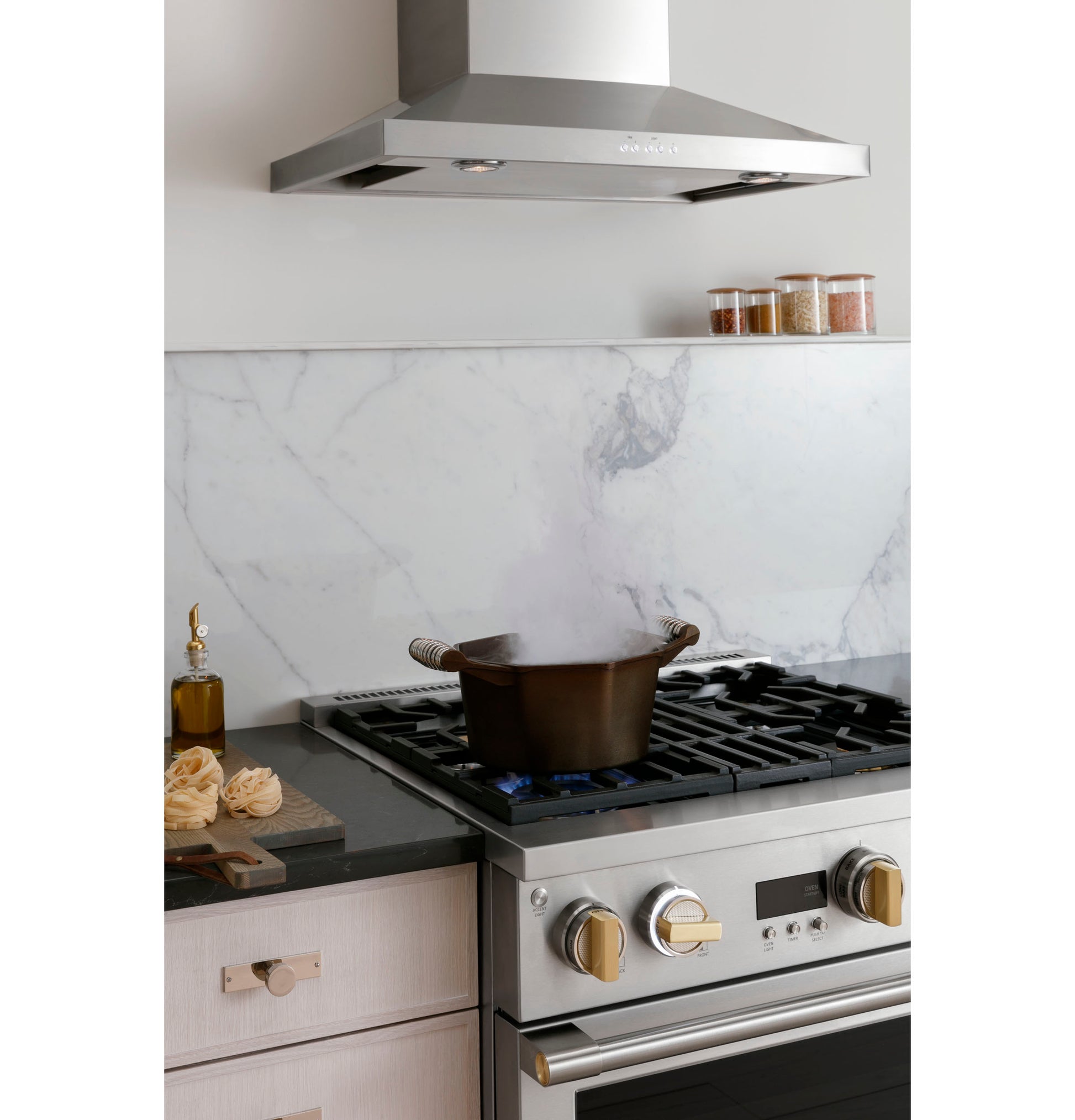 Stainless steel wall-mounted vent hood above gas range with pot steaming and marble backsplash in modern kitchen