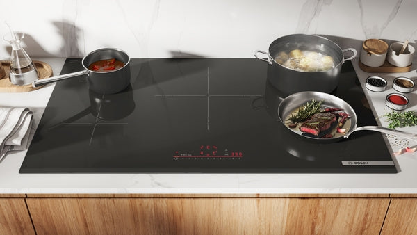 Modern black induction cooktop with four active cooking zones preparing pasta, sauce, and steak in kitchenware collection