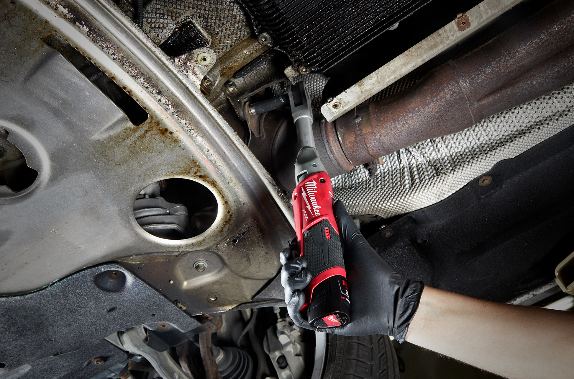 Hand in black glove holding red Milwaukee M12 FUEL extended reach ratchet tightening bolts under vehicle for repairs