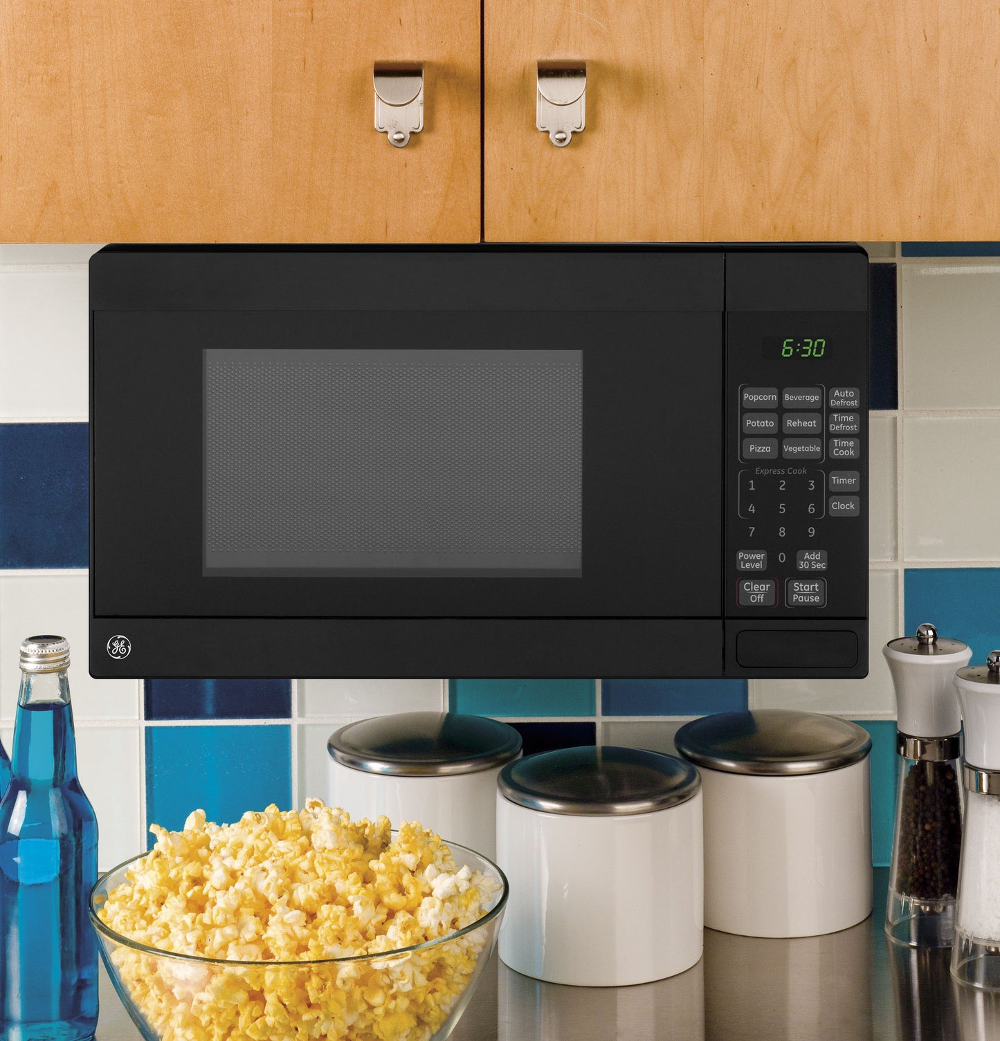 Black undercabinet microwave with digital control panel mounted under wood cabinets above kitchen counter with popcorn bowl and containers