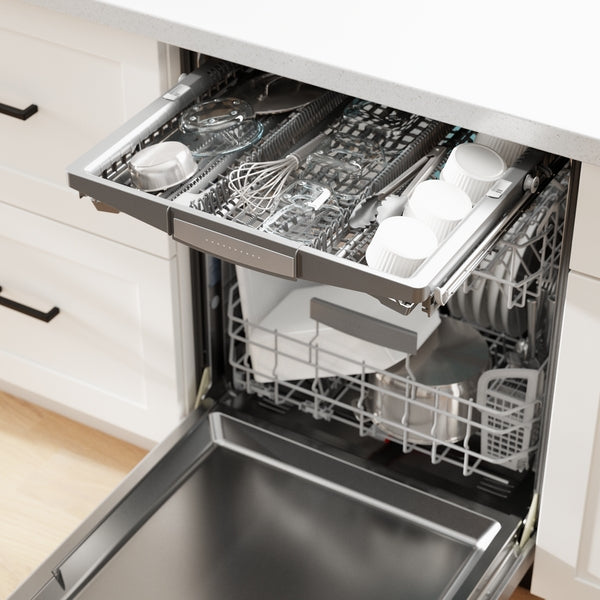Open stainless steel dishwasher with upper tray holding cups and kitchen utensils inside modern white cabinetry