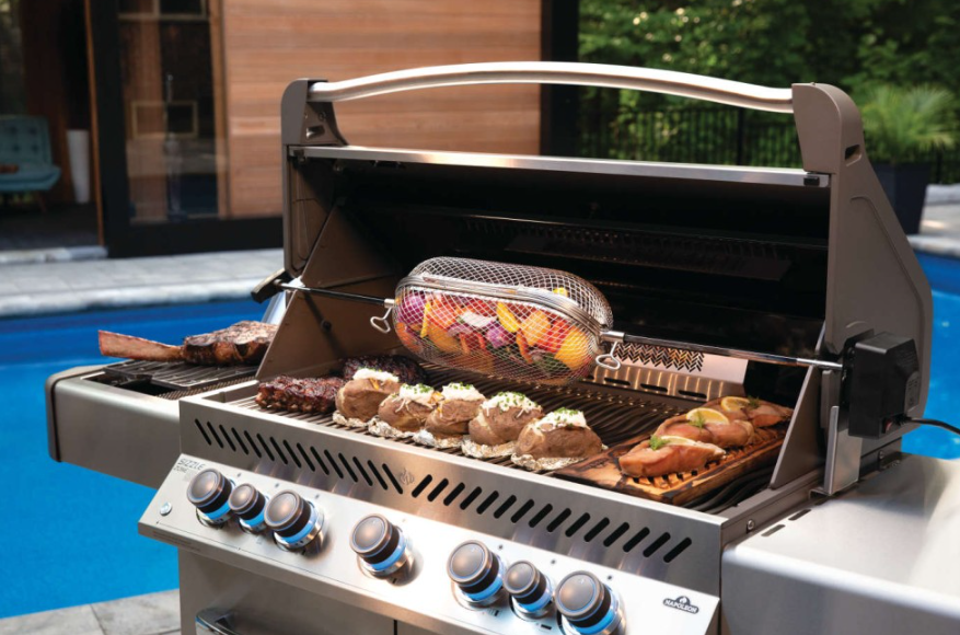 Outdoor stainless steel grill with rotisserie basket cooking mixed vegetables and grilled meats, ideal for grilling accessories and outdoor living.