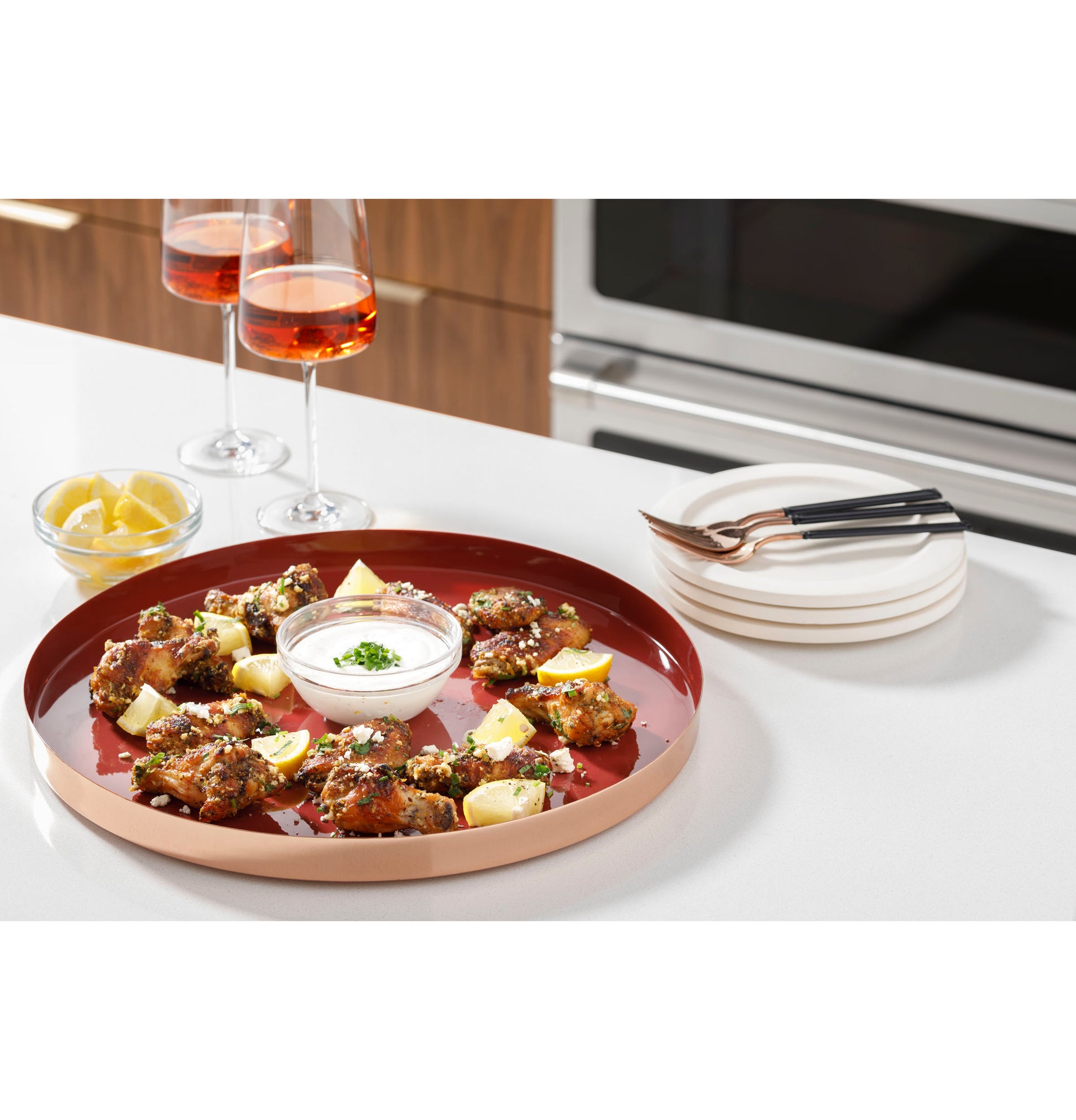 Round serving tray with chicken bites, lemon wedges, dipping sauce, and two glasses of rosé on kitchen counter