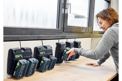 Person placing a 18V battery into a black charger with multiple Festool battery chargers lined up on a workbench
