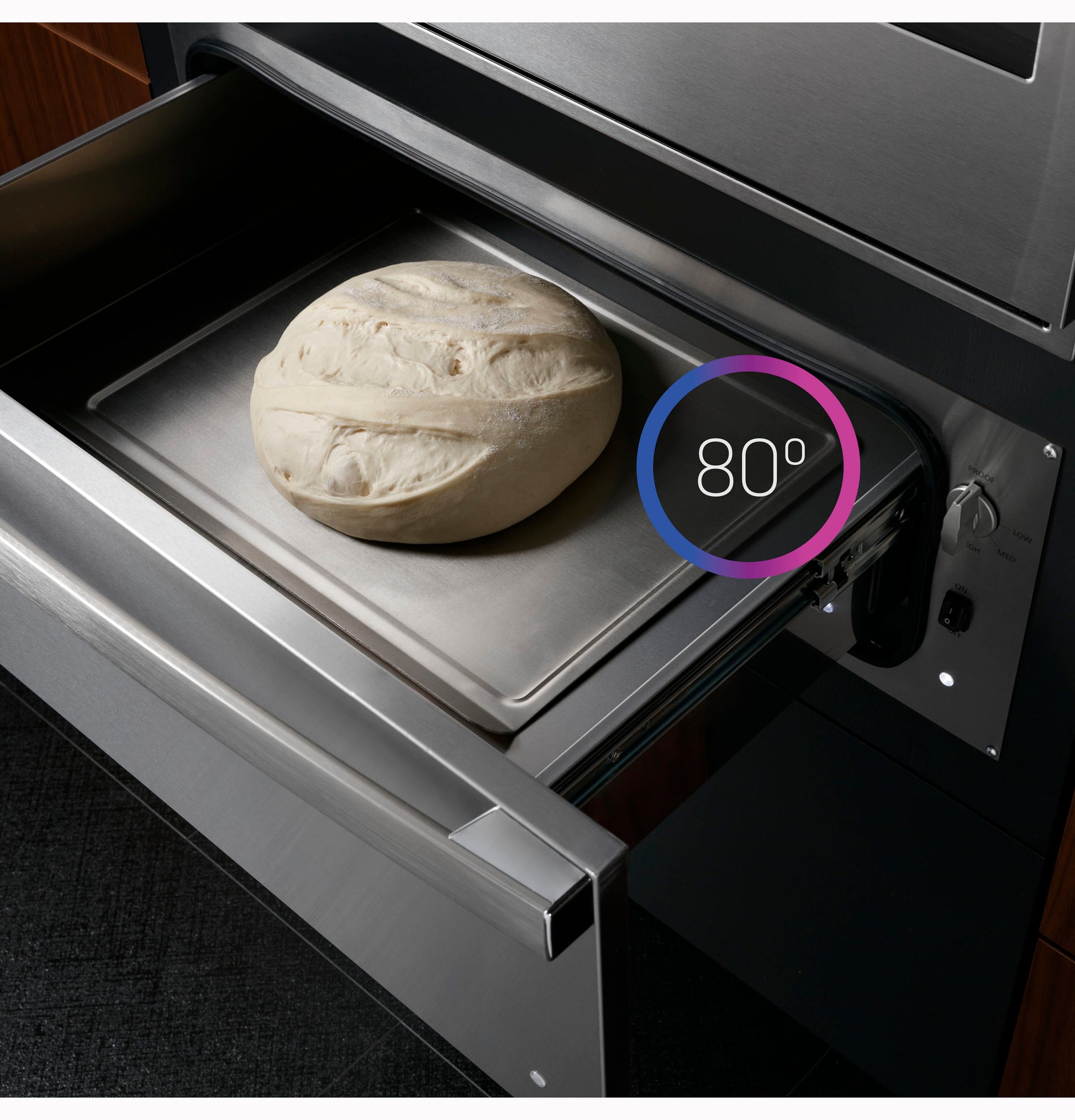 Open stainless steel warming drawer with bread dough on tray set at 80 degrees in modern kitchen appliances collection