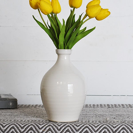 White ceramic vase with tear drop shape holding bright yellow tulips on patterned cloth surface