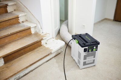 Compact dust extraction unit with flexible white air supply hose positioned near wooden staircase during home renovation